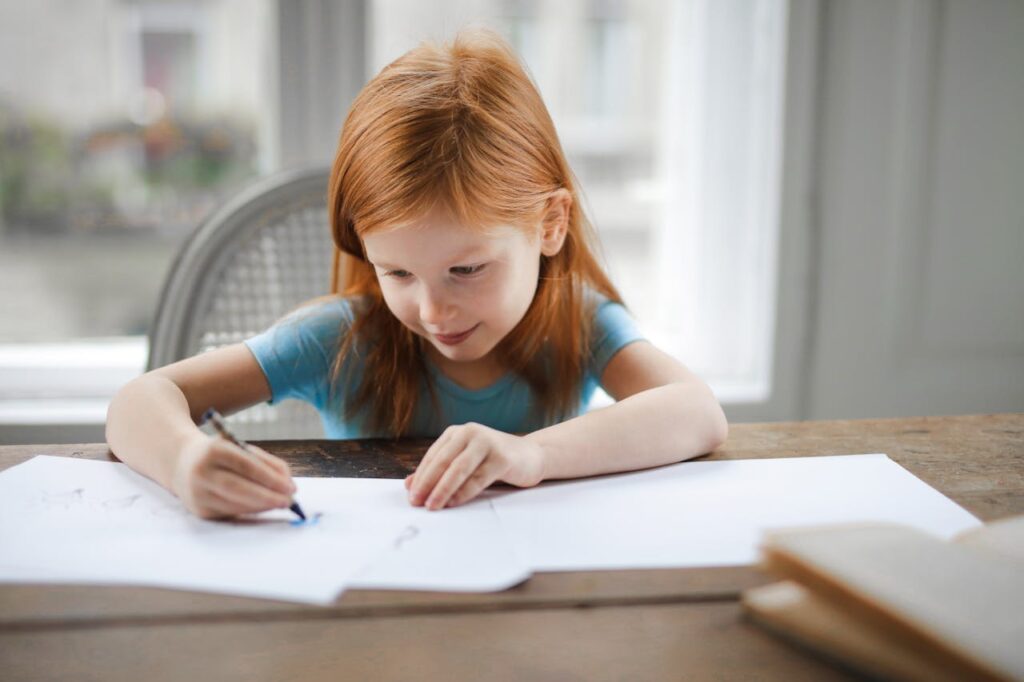 A young girl with red hair focused on drawing at a wooden desk indoors.